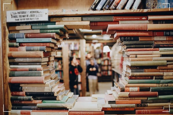 A pile of books one on top of the other in such a way that there is a circle shaped opening in the middle that allows to see the library and the people in the background.